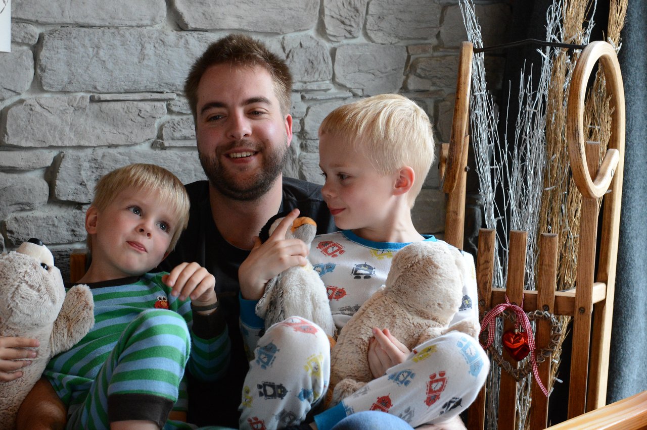 A man sits with two young children in pajamas, both holding stuffed animals and smiling.
