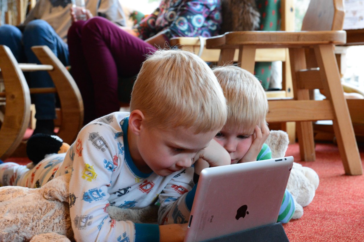 Two young children in pajamas lie on the floor, closely watching a tablet screen together.