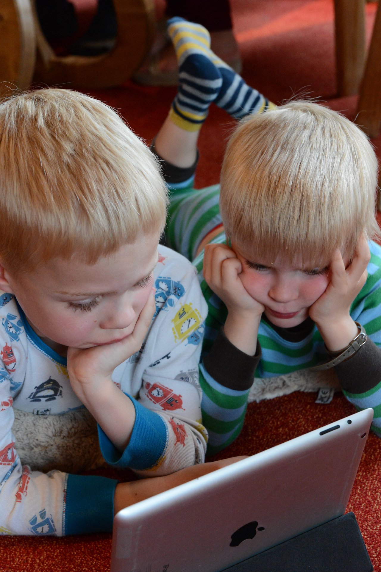 Two young children in pajamas lie on the floor, resting their heads on their hands, watching a tablet screen.