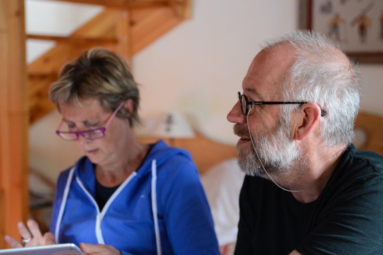A man with glasses and a beard talks while a woman in a blue hoodie focuses on a tablet.