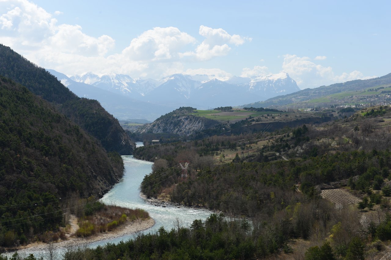A winding river flows through a green valley, with snow-capped mountains in the background under a partly cloudy sky.