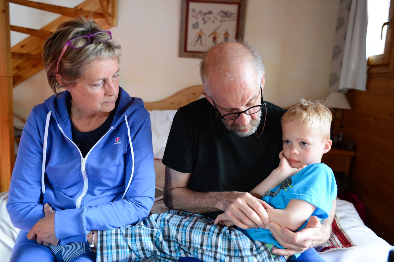 An older man holds a young boy on his lap while an older woman sits beside them, looking concerned.