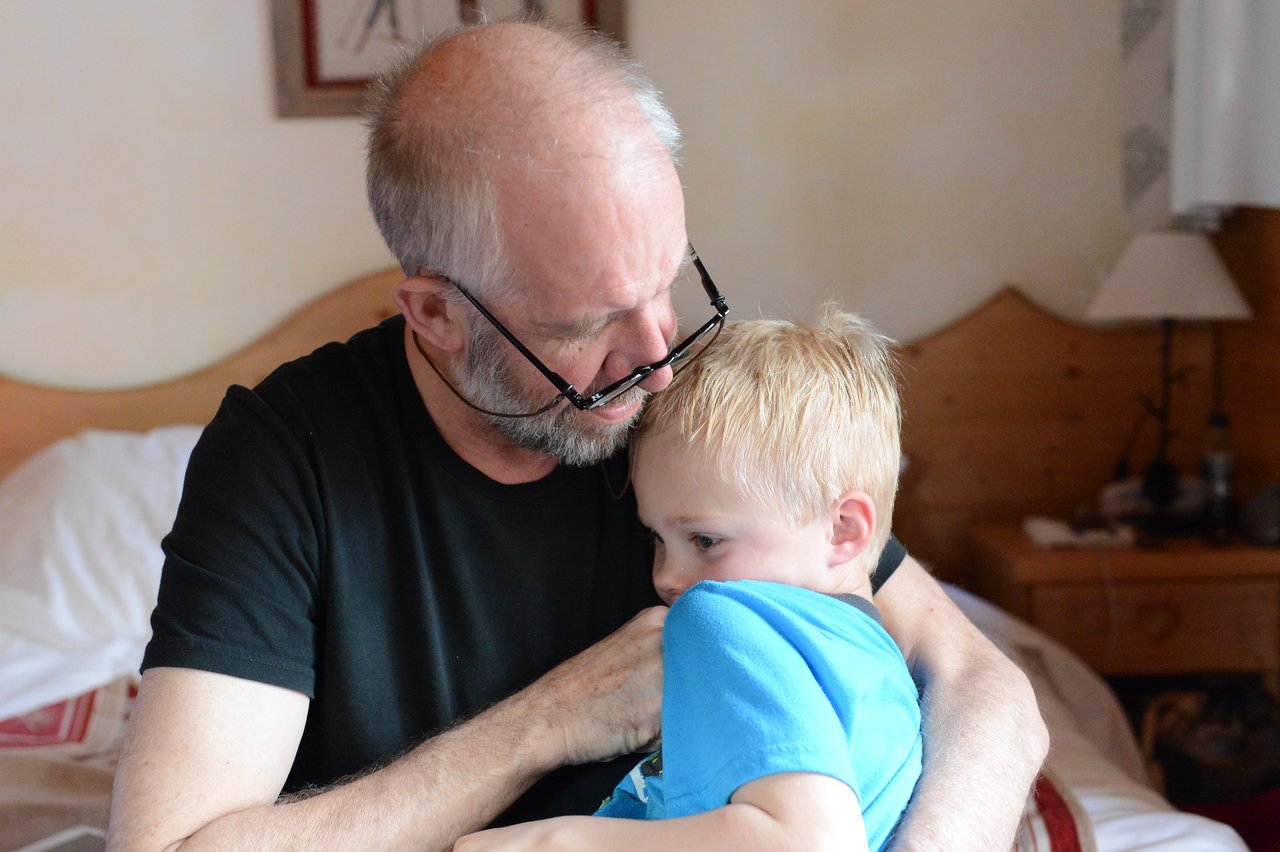 An older man in a black shirt hugs a young boy in blue, comforting him in a bedroom.