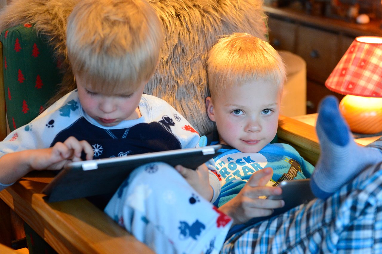 Two young children in pajamas sit closely in a chair, each focused on a tablet screen.