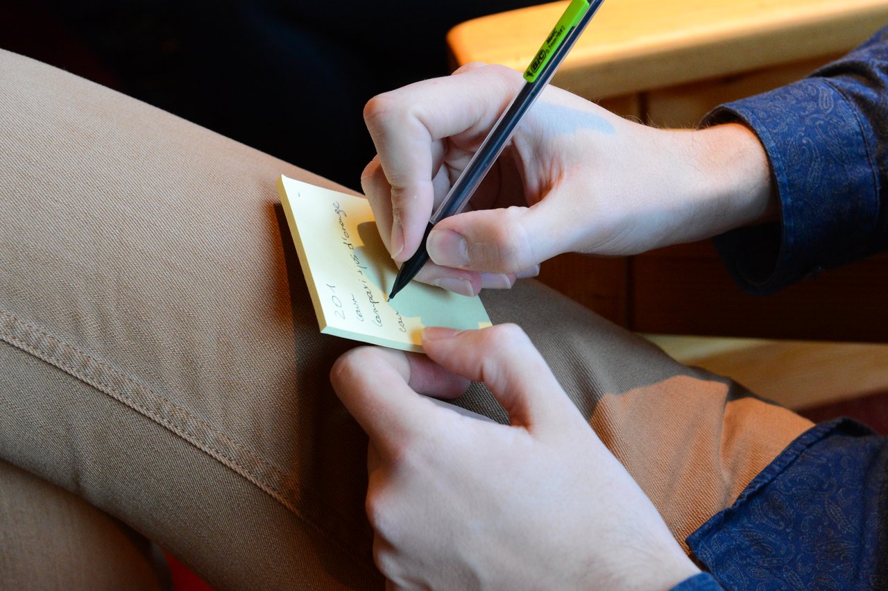 A person writes on a small sticky note with a black pen while resting it on their lap.