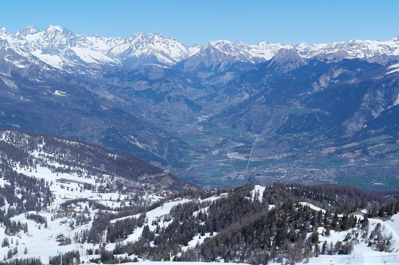 Snow-covered mountain slopes with a small village, overlooking a valley with distant peaks under a clear blue sky.