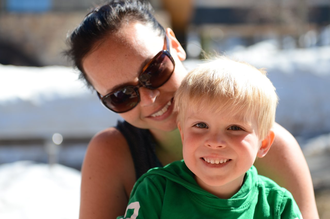 A smiling woman wearing sunglasses leans close to a happy young boy in a green shirt outdoors.