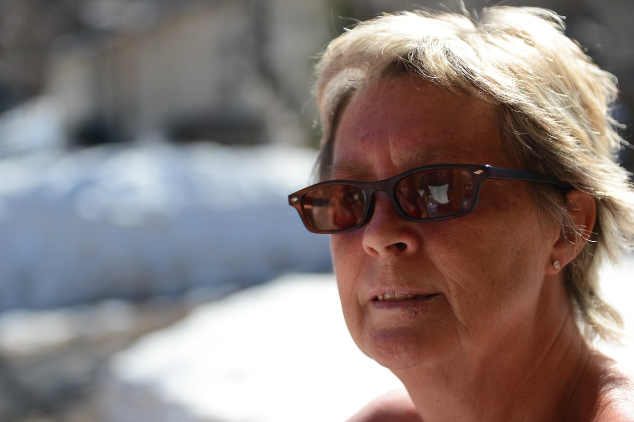 A woman wearing sunglasses looks slightly away from the camera, with a snowy background behind her.