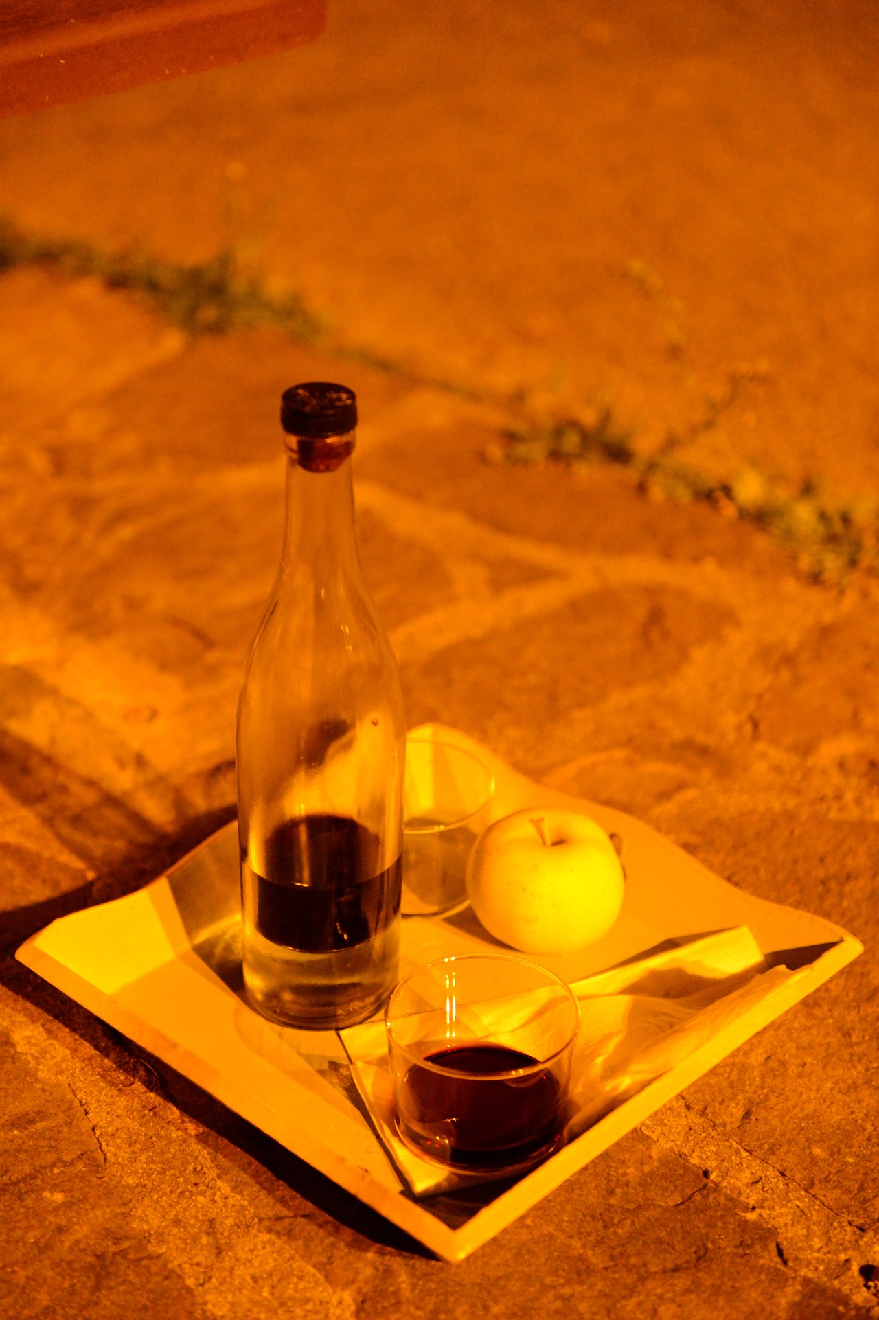 A tray with a bottle of dark liquid, two glasses, an apple, and a napkin on a stone surface.