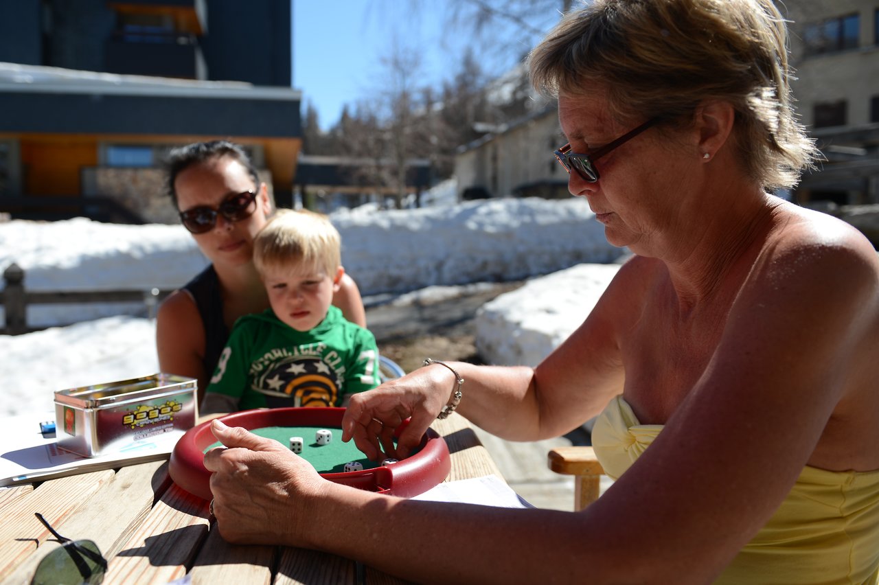 A woman in sunglasses plays a dice game at an outdoor table, while a child watches with interest.