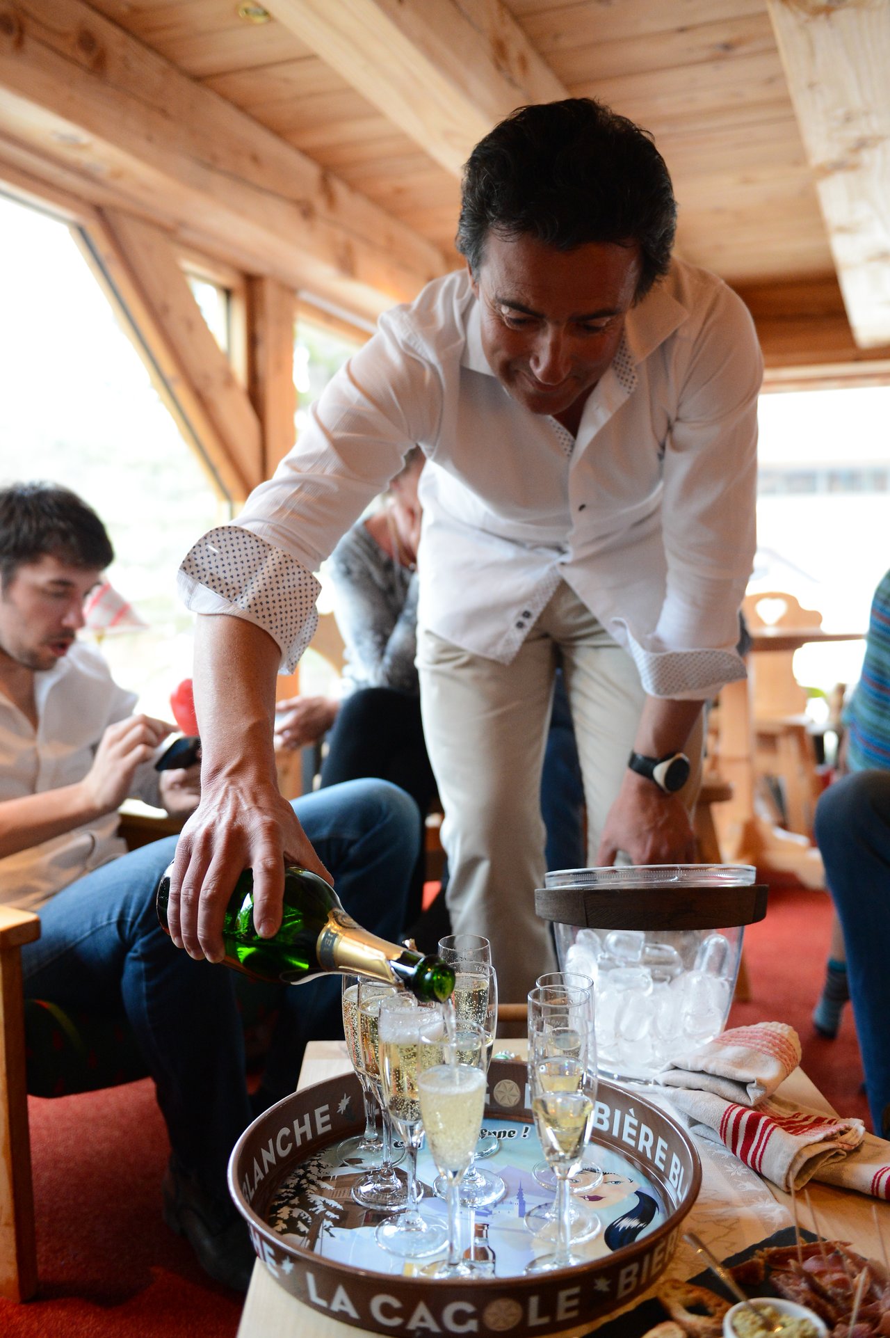 A man in a white shirt pours champagne into glasses on a tray, with people sitting nearby.