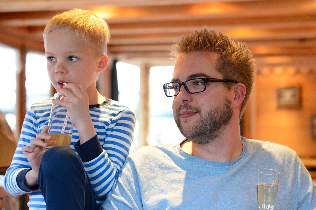 A young boy drinks juice through a straw while sitting next to a man holding a glass of champagne.