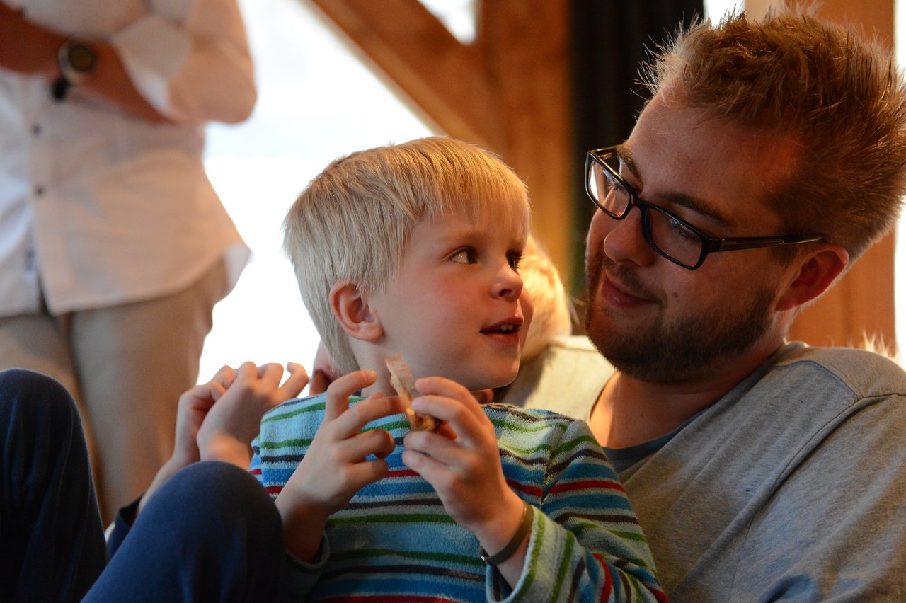 A man with glasses smiles while holding a young child in a striped shirt, who is eating a snack.