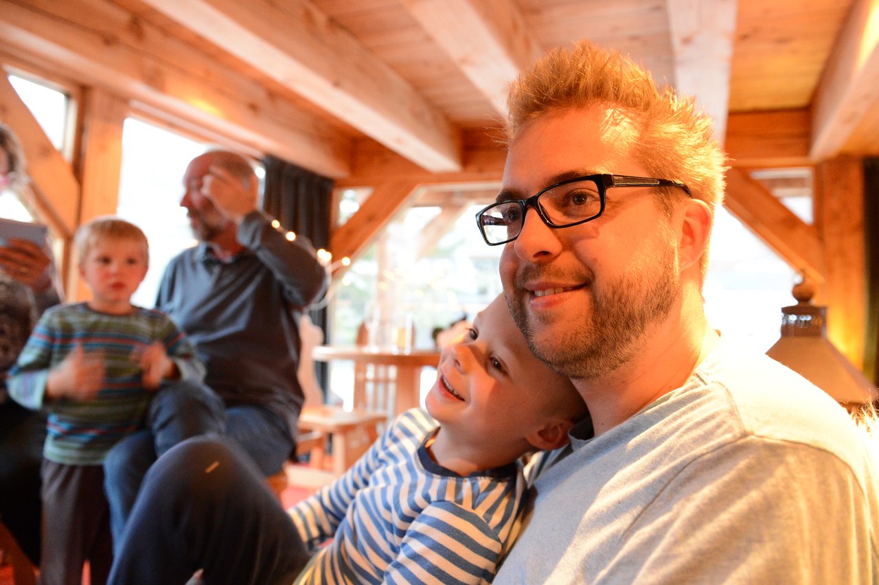 A man with glasses smiles while a young child leans on his shoulder, both looking happy.