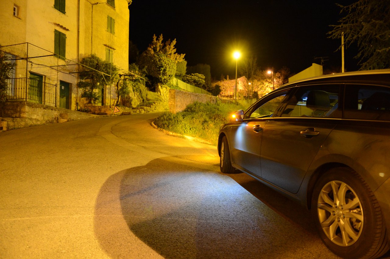A car with headlights on is parked on a curved road at night near a building with green shutters.