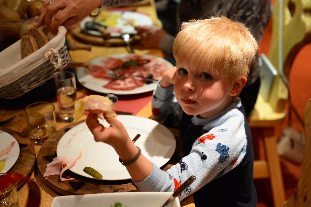A young child in pajamas sits at a dinner table, holding food while looking towards the camera.