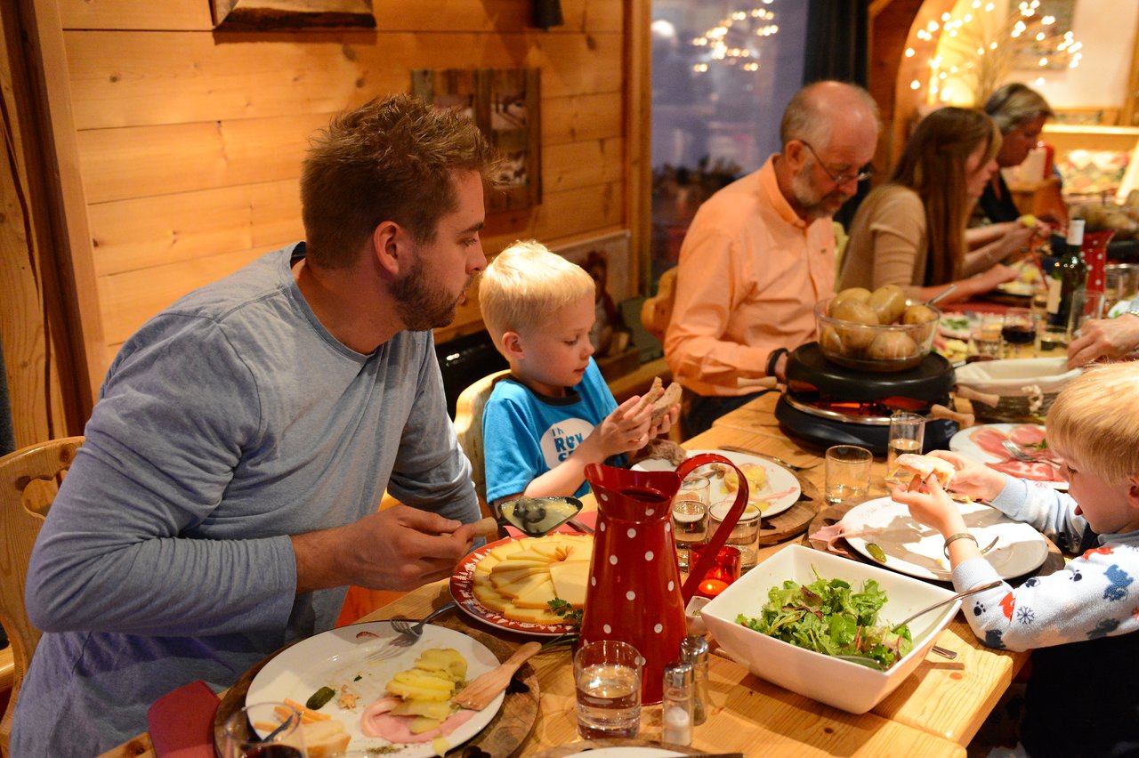 A family gathers around a wooden table, sharing a meal with cheese, bread, and salad in a cozy setting.