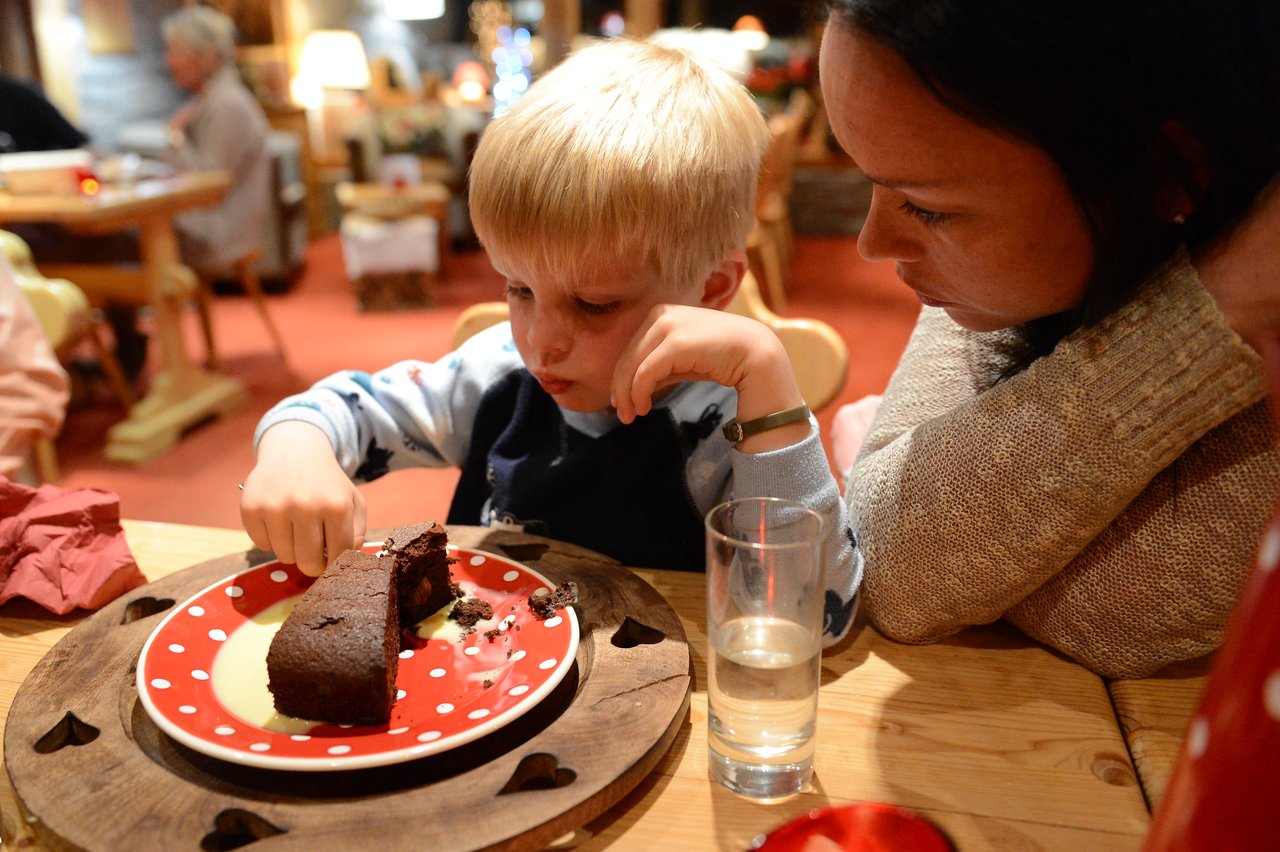 A young child eats chocolate cake while an adult watches closely at a wooden table in a cozy setting.