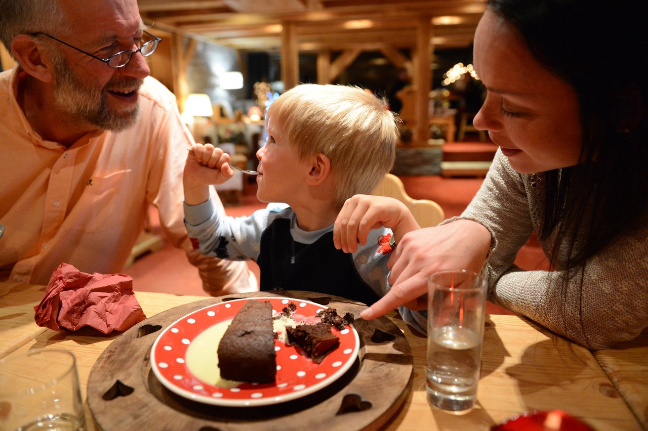 A young child eats chocolate cake while sitting between two smiling adults at a wooden table.