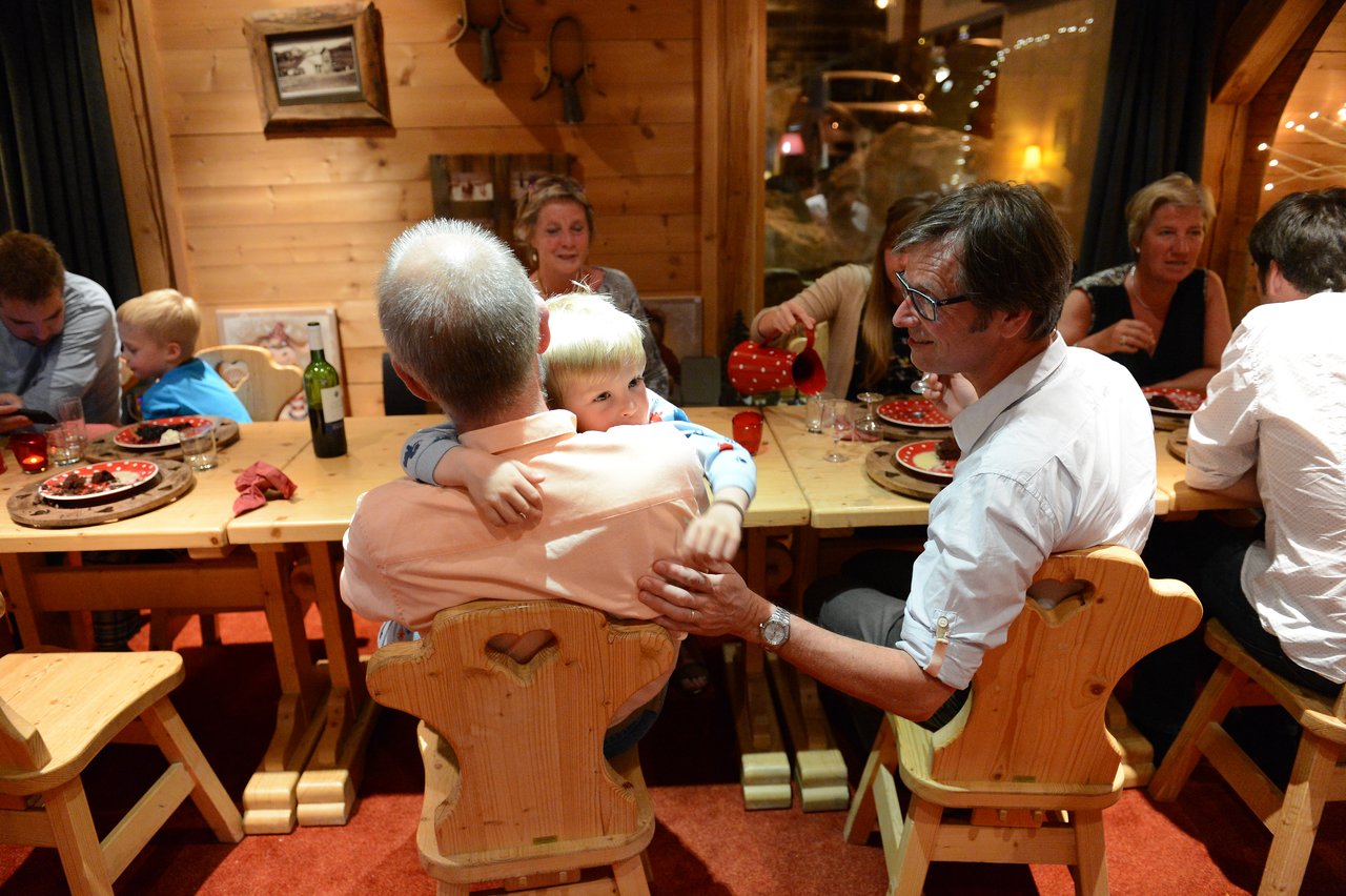 A child hugs an older man from behind while seated at a wooden dining table with family and friends.