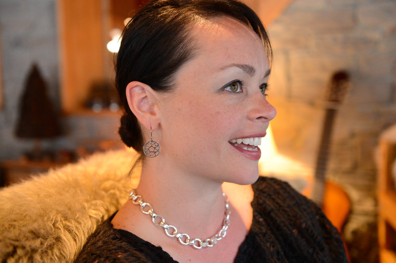 A woman with dark hair smiles while looking to the side, wearing a silver necklace and dangling earrings.