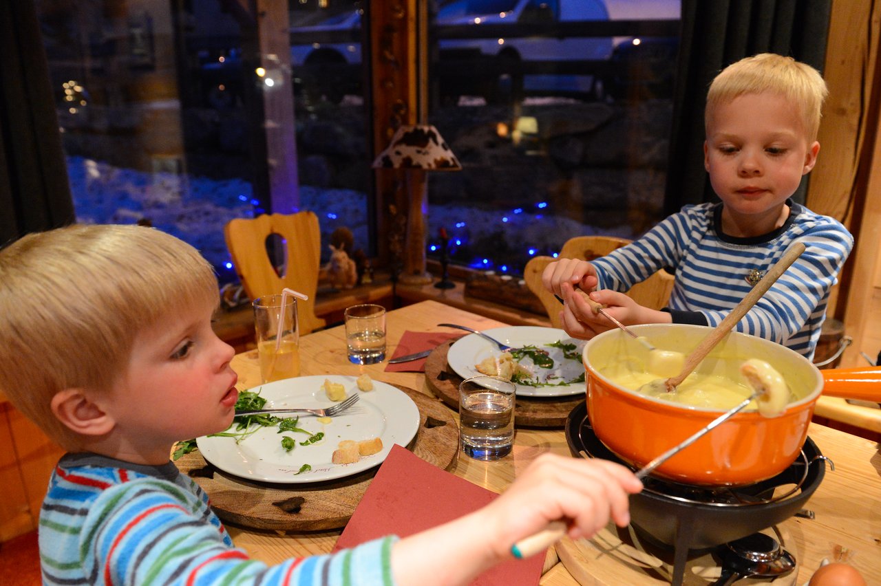 Two children in striped shirts dip bread into a pot of cheese fondue at a wooden dining table.