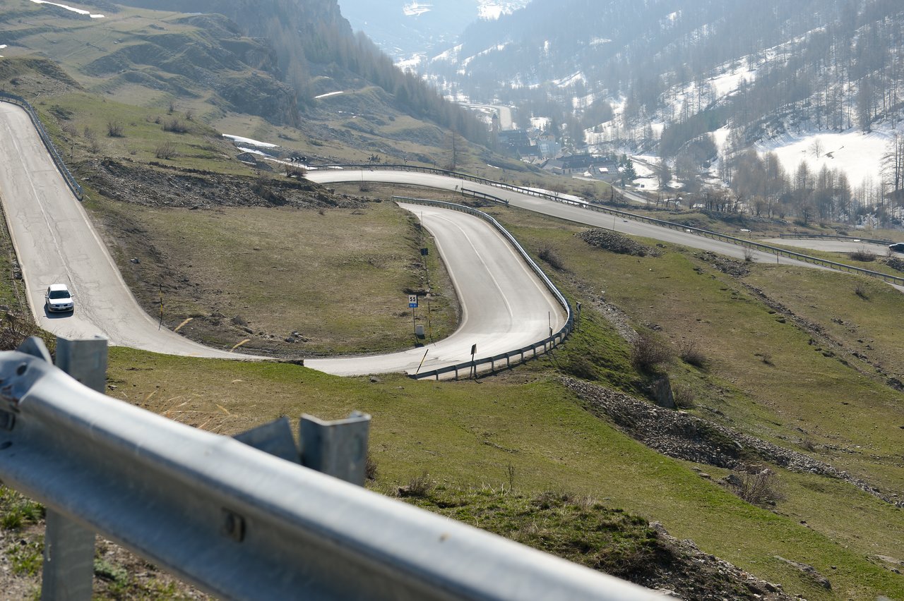 A winding mountain road with a white car driving downhill, surrounded by grassy slopes and patches of snow.