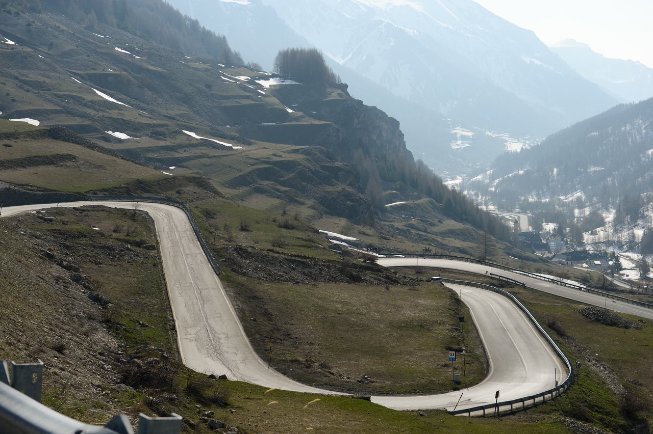 A winding mountain road with sharp curves, surrounded by grassy slopes and patches of snow.