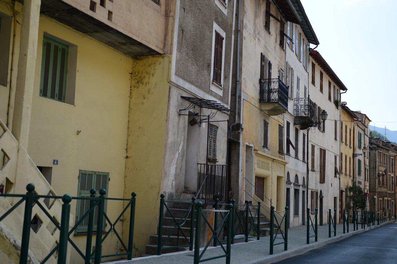 A row of old buildings with balconies and shutters lines a quiet street with green railings.