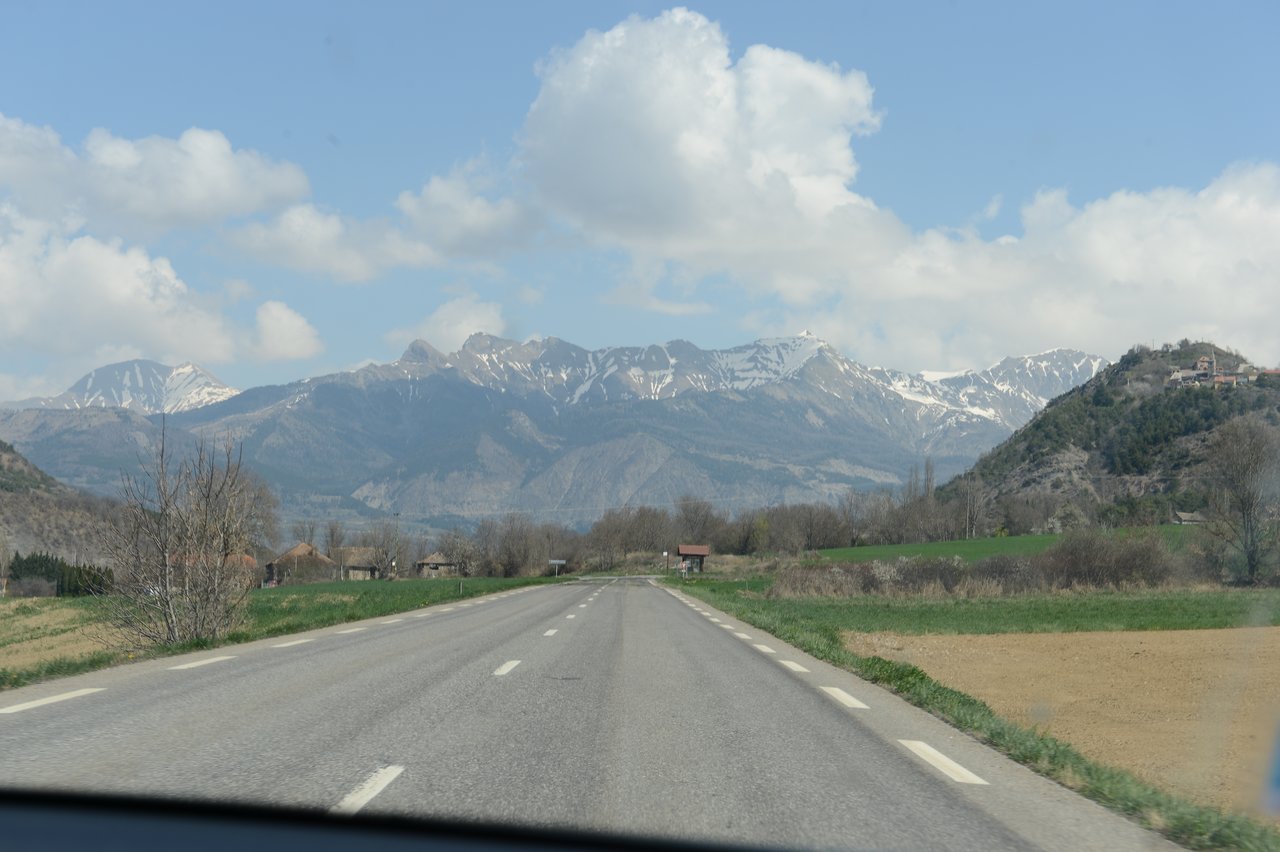 A two-lane road leads toward snow-capped mountains under a partly cloudy sky.