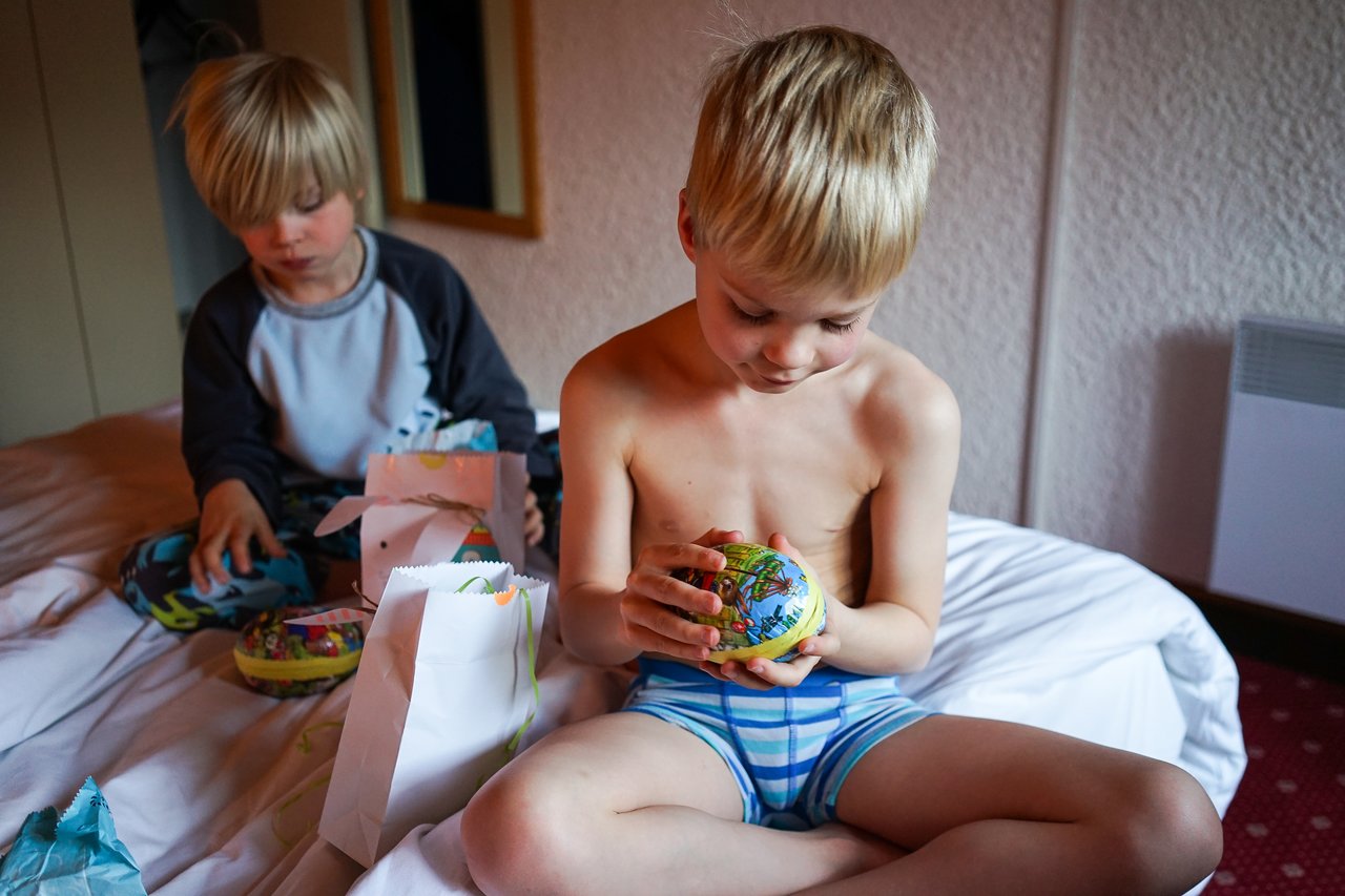 Two children sit on a bed, opening gift bags and examining colorful candy-filled eggs.