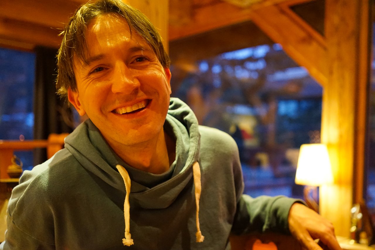 A man in a blue hoodie smiles while leaning on a wooden surface in a warmly lit indoor setting.
