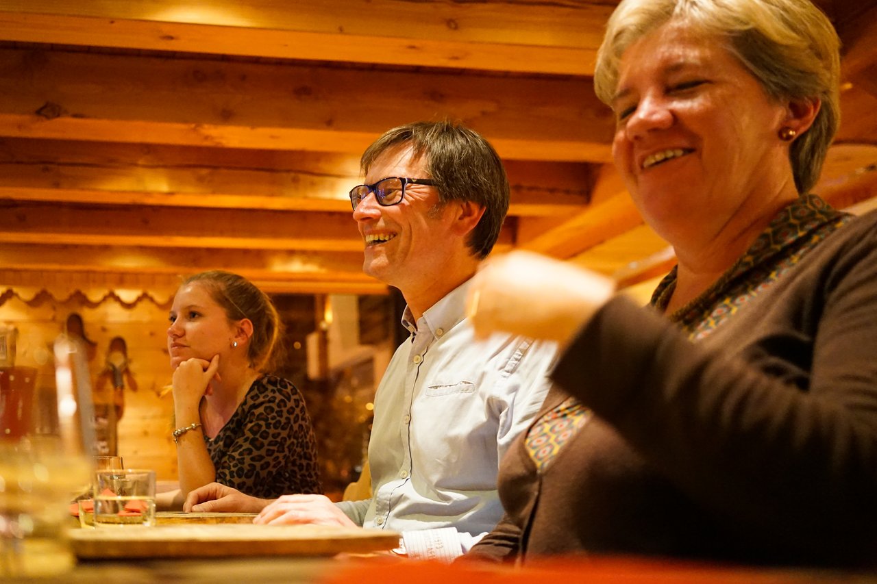 Three people sit at a wooden table, smiling and engaged in conversation in a warmly lit setting.