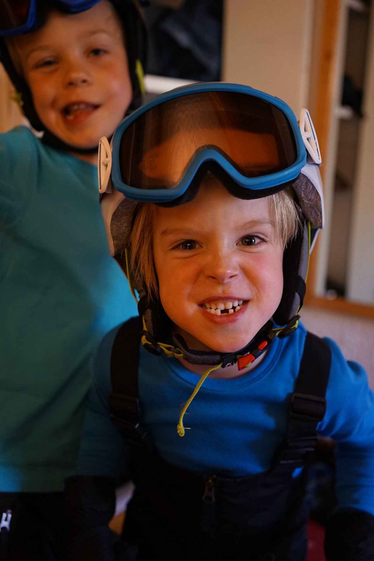 Two children in ski gear smile playfully, wearing helmets and goggles indoors.