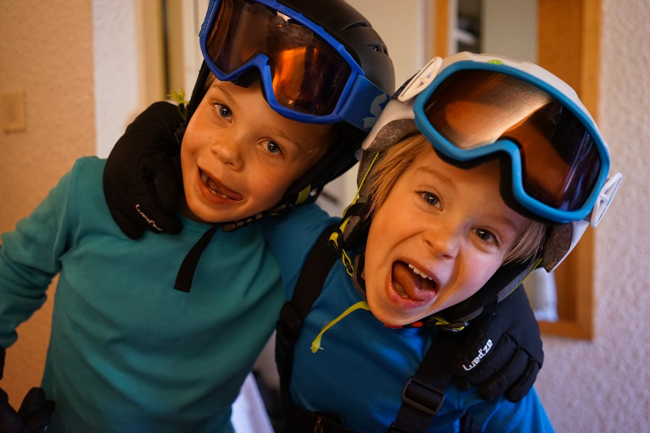 Two children in ski gear smile and pose playfully indoors, wearing helmets, goggles, and gloves.