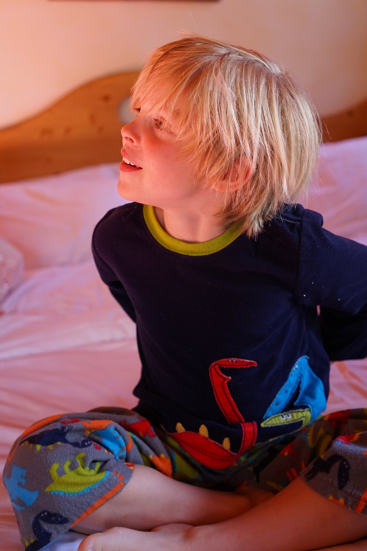 A child with blonde hair sits on a bed, wearing colorful dinosaur pajamas, looking to the side and smiling.