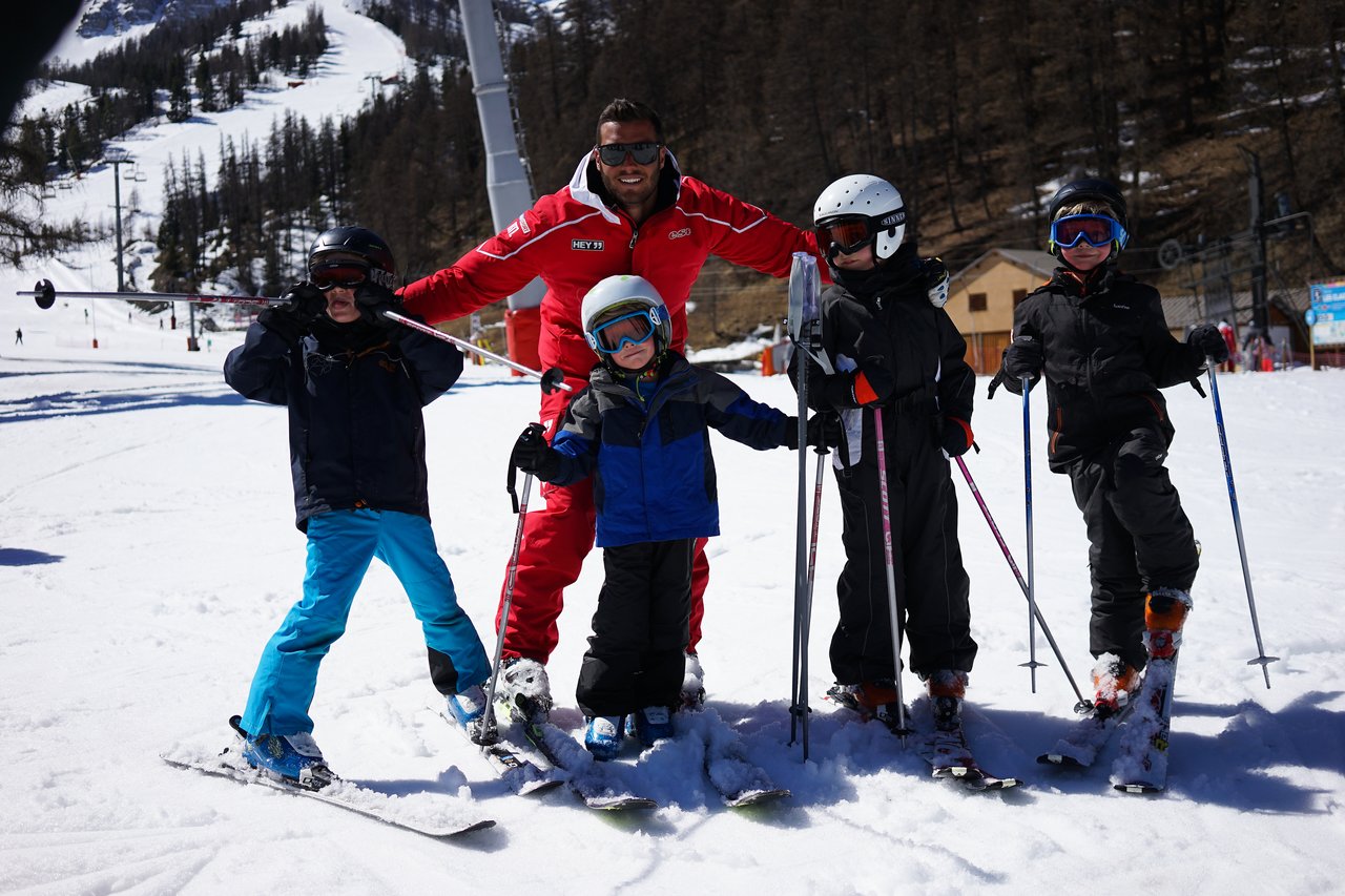 A ski instructor in a red suit poses with four children in ski gear on a snowy slope.
