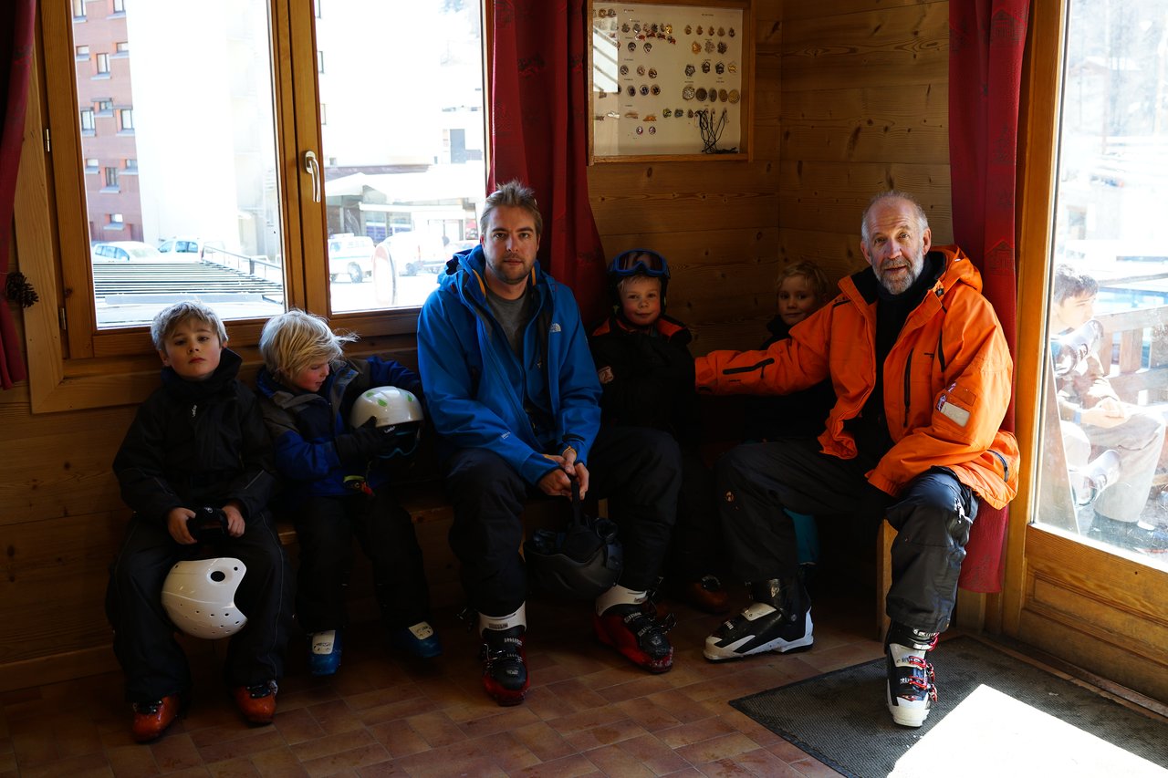 A group of adults and children in ski gear sit on a bench inside a wooden cabin, holding helmets.