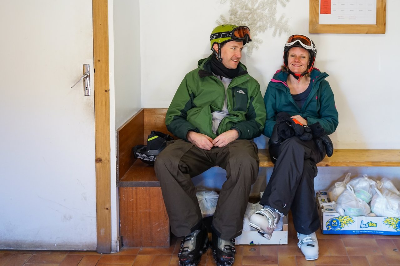 Two people in ski gear sit on a bench indoors, smiling and talking.