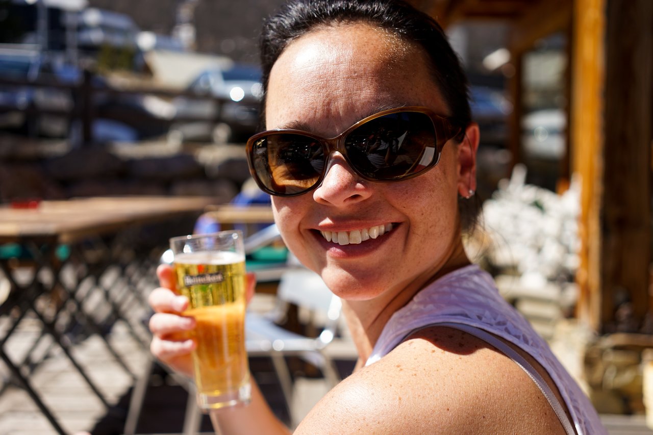 A woman wearing sunglasses smiles while holding a glass of beer outdoors.