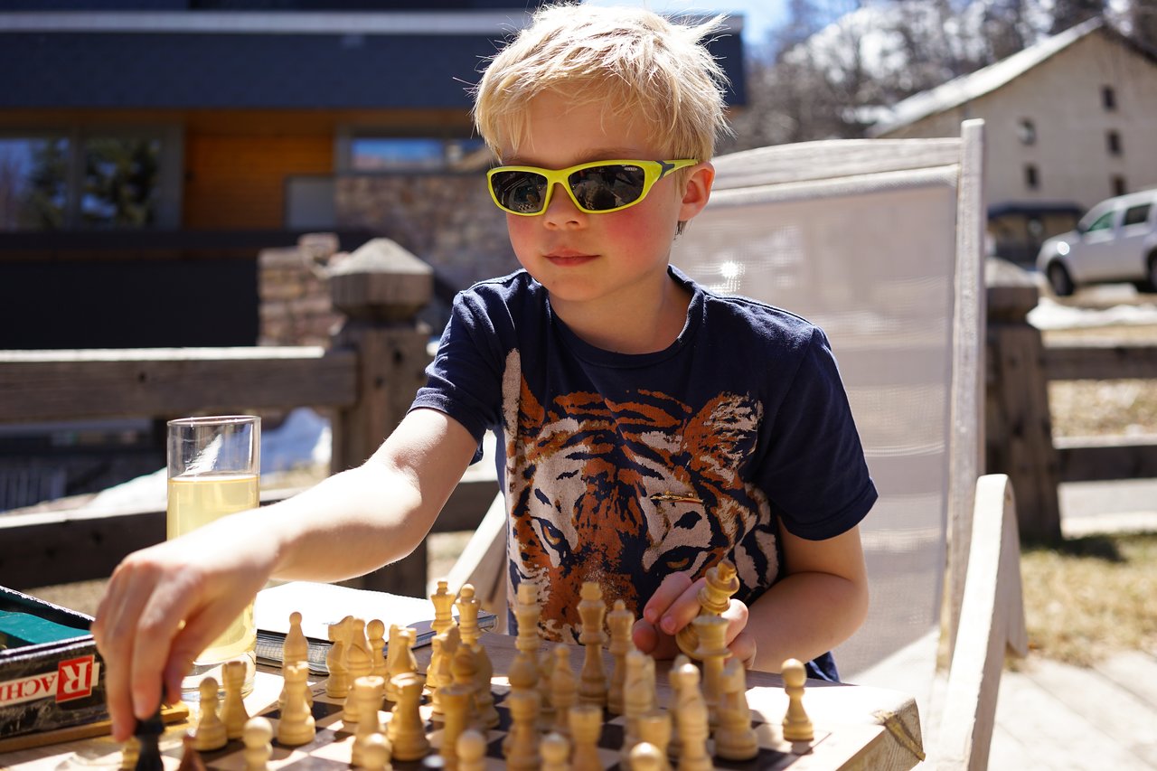 A child wearing sunglasses plays chess outdoors, reaching to move a piece.