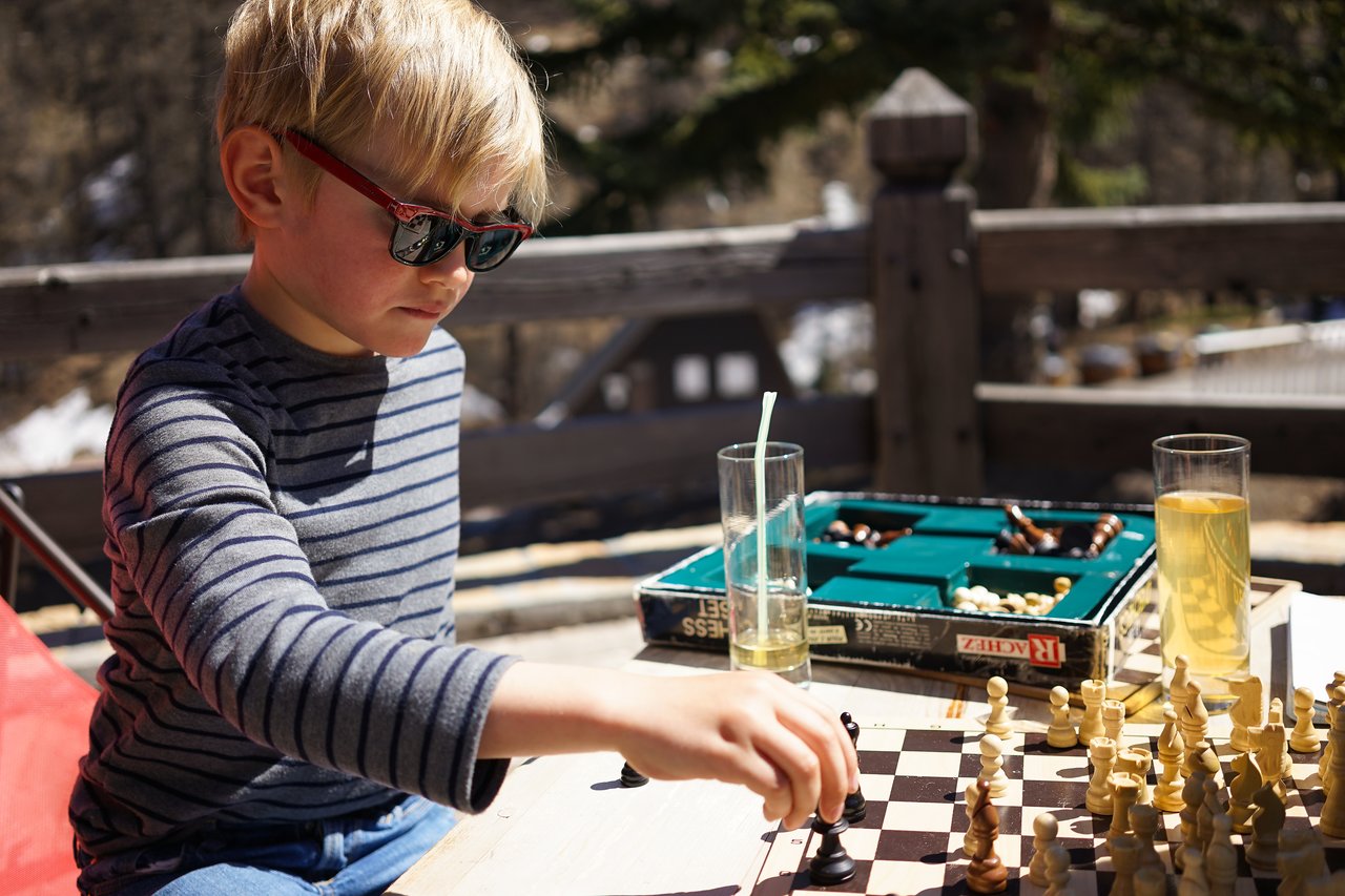 A child wearing sunglasses plays chess outdoors, moving a piece on the board with focus.