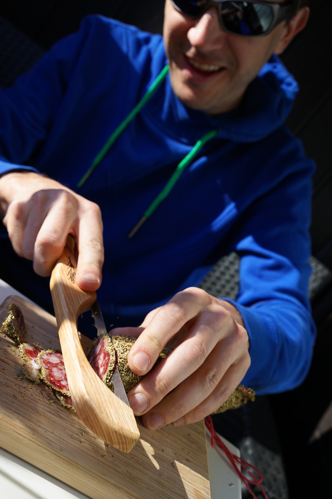 A person in a blue hoodie slices a herb-crusted sausage on a wooden cutting board using a special knife.
