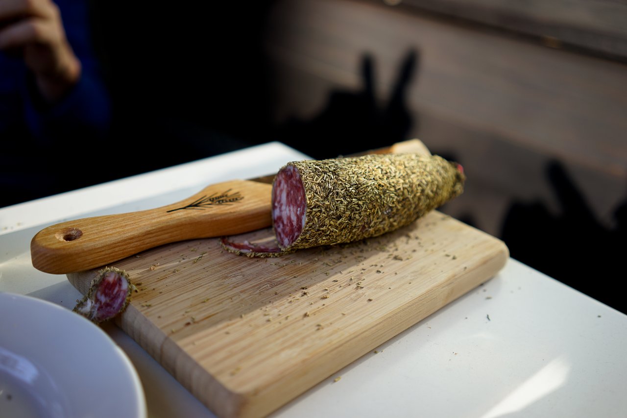 A partially sliced herb-crusted salami on a wooden cutting board with a knife resting beside it.
