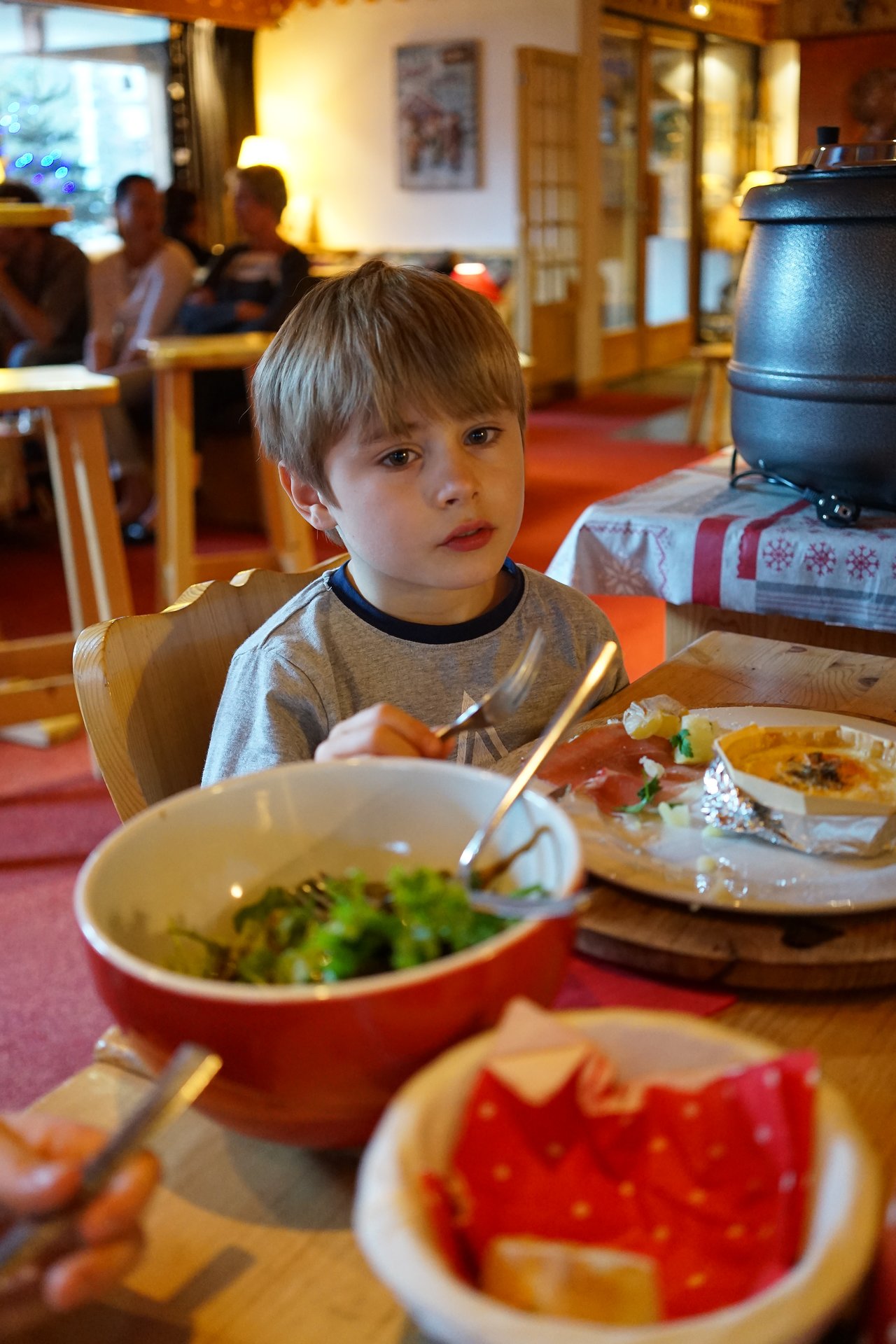 A young boy sits at a restaurant table, holding a fork while eating a meal with salad and bread.
