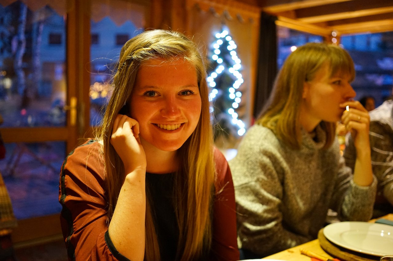 A smiling woman sits at a table in a warmly lit restaurant, with another person eating beside her.