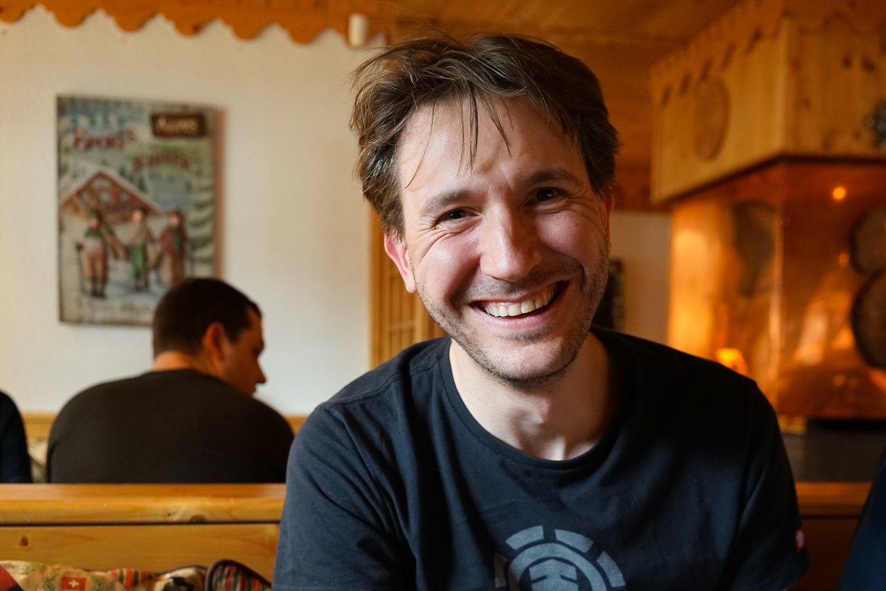 A man with short brown hair and a black shirt smiles at the camera in a cozy indoor setting.