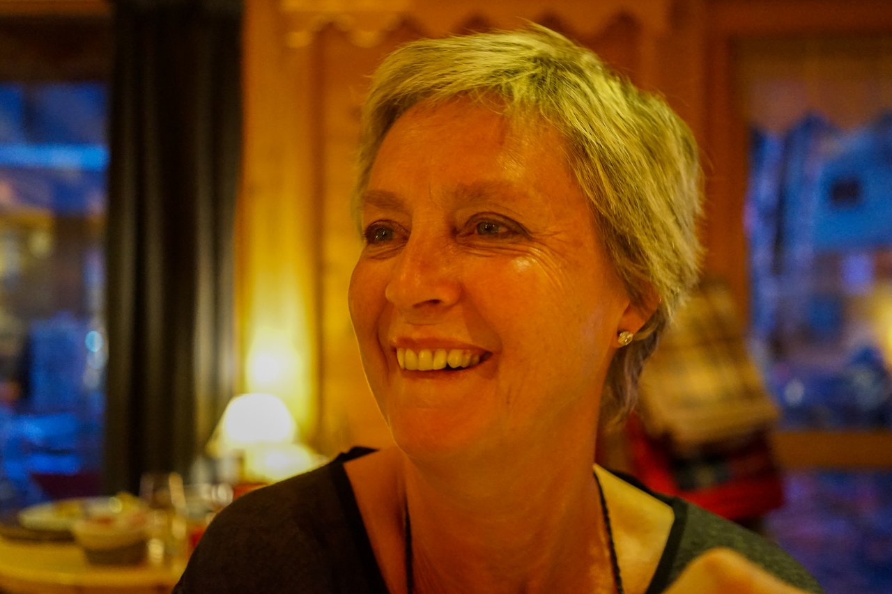 A woman with short hair smiles warmly in a dimly lit indoor setting.