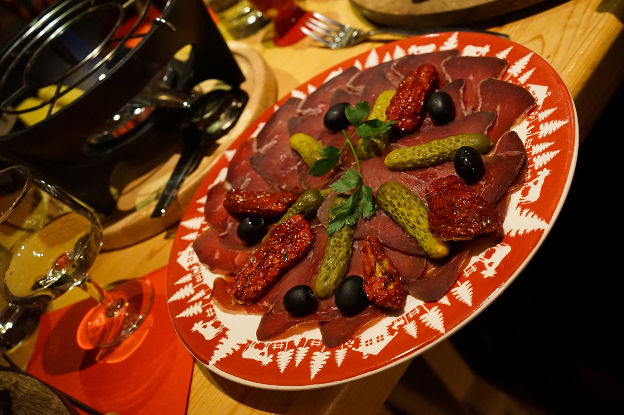 A plate of cured meat with pickles, sun-dried tomatoes, black olives, and parsley, served on a wooden table.