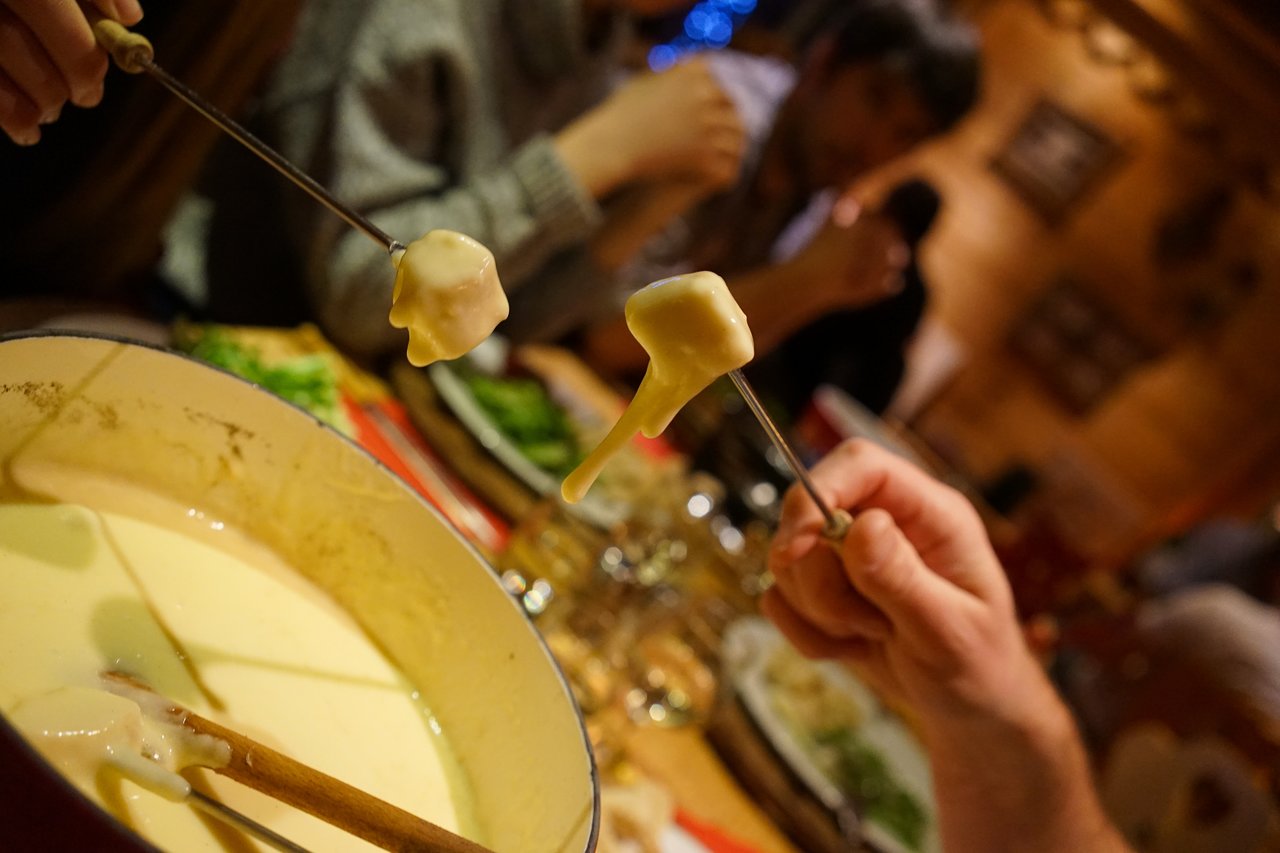 Two hands hold fondue forks with bread dipped in melted cheese from a pot at a dining table.